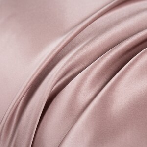 May include: Close-up of a soft, smooth, and silky fabric in a light dusty rose color. The fabric is draped and folded, creating a luxurious and elegant look.