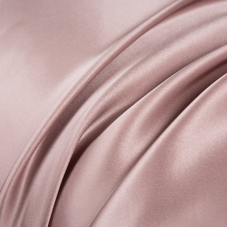May include: Close-up of a soft, smooth, and silky fabric in a light dusty rose color. The fabric is draped and folded, creating a luxurious and elegant look.