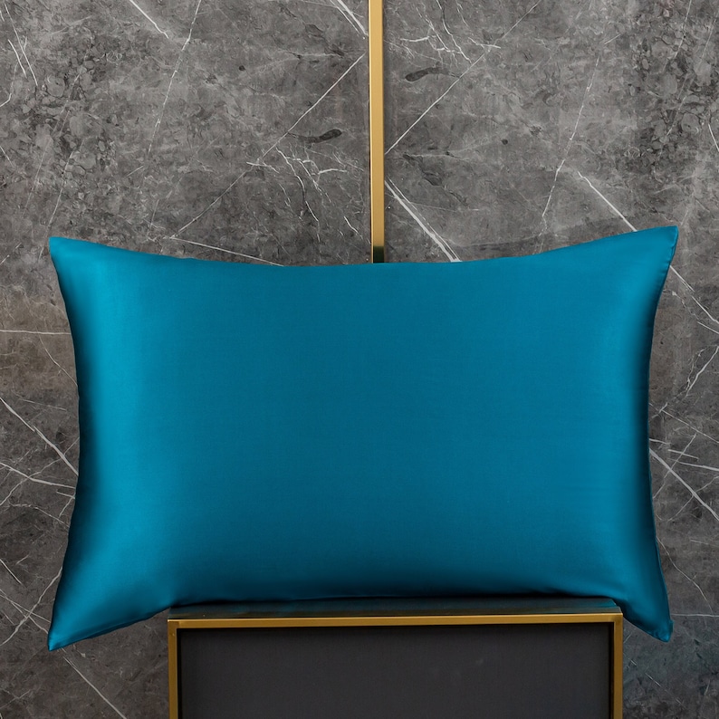 May include: A teal blue satin pillowcase, shown on a black and gold stand against a gray marble background.