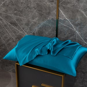 May include: A teal blue satin pillowcase with a smooth, silky texture. The pillowcase is folded and draped over a dark gray surface.