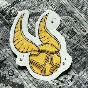 May include: A die-cut sticker of a golden Snitch with black outlines and white borders. The Snitch has two wings and a round body, with star accents. The sticker is on a newspaper background.