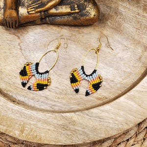 Könnte beinhalten: Ein Paar handgefertigte Makramee-Ohrringe. Die Ohrringe haben einen goldfarbenen ovalen Reifen mit einem farbenfrohen gewebten Design in Schwarz, Gelb, Hellblau und Pfirsich. Sie werden auf einer Holzoberfläche präsentiert.
