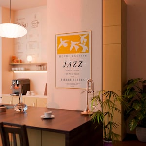 Könnte beinhalten: Gerahmtes Poster mit dem Text "JAZZ" und "HENRI MATISSE" in einem Café. Das Poster hat ein gelbes und weißes Design. Eine Trompete und eine Pflanze sind ebenfalls zu sehen.