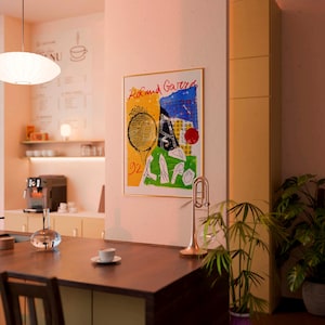 May include: A framed art print with the words "Roland Garros" in red, featuring a tennis racket, and abstract shapes in yellow, green, and blue. A gold trumpet and a potted plant are in the foreground. The setting appears to be a cafe.