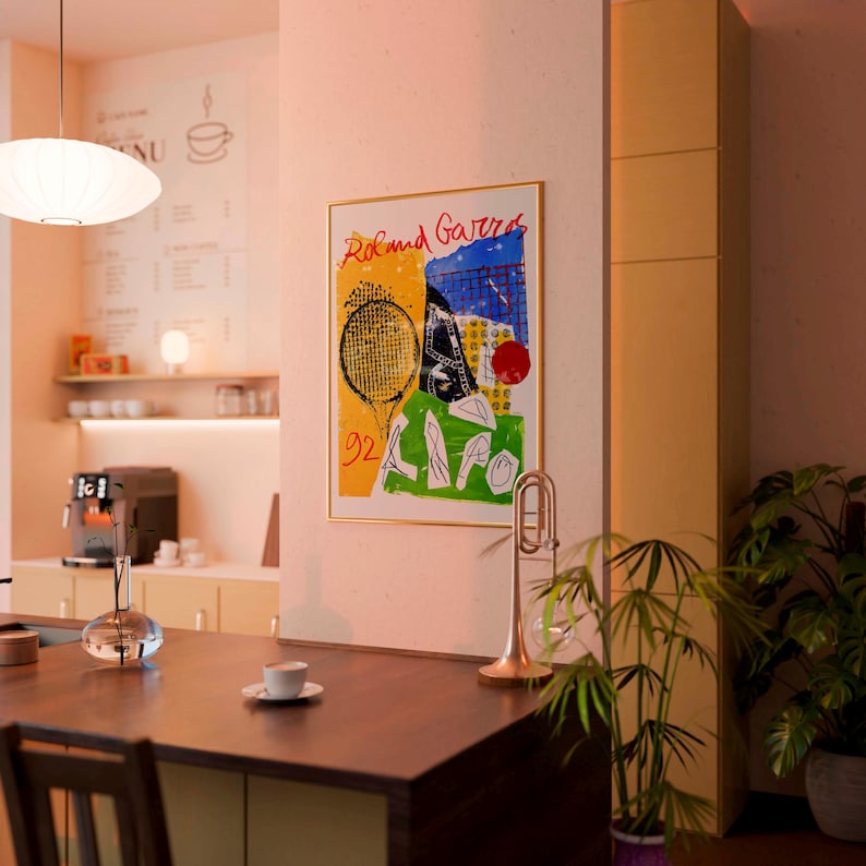 May include: A framed art print with the words "Roland Garros" in red, featuring a tennis racket, and abstract shapes in yellow, green, and blue. A gold trumpet and a potted plant are in the foreground. The setting appears to be a cafe.