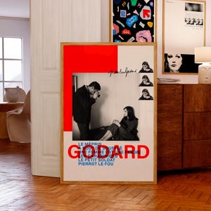 Op de afbeelding: Een ingelijste poster met een rode en witte achtergrond, met een zwart-wit afbeelding van een man en een vrouw. Het woord "GODARD" staat in grote rode letters. Andere ingelijste kunstwerken zijn zichtbaar.
