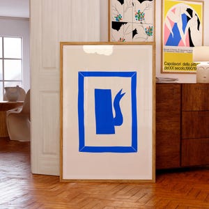 Pode incluir: Uma grande impressão de arte emoldurada com um design abstrato azul arrojado sobre um fundo creme. A obra de arte está em uma moldura de madeira clara e está encostada em uma parede. Obras de arte emolduradas adicionais são visíveis no fundo, juntamente com um armário de madeira e uma porta branca.