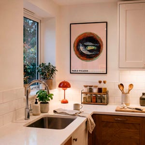 May include: A kitchen scene with a sink, countertop, and cabinets. A framed Pablo Picasso print hangs on the wall. A small orange lamp sits on the counter, along with spice jars, wooden spoons, and a cutting board.