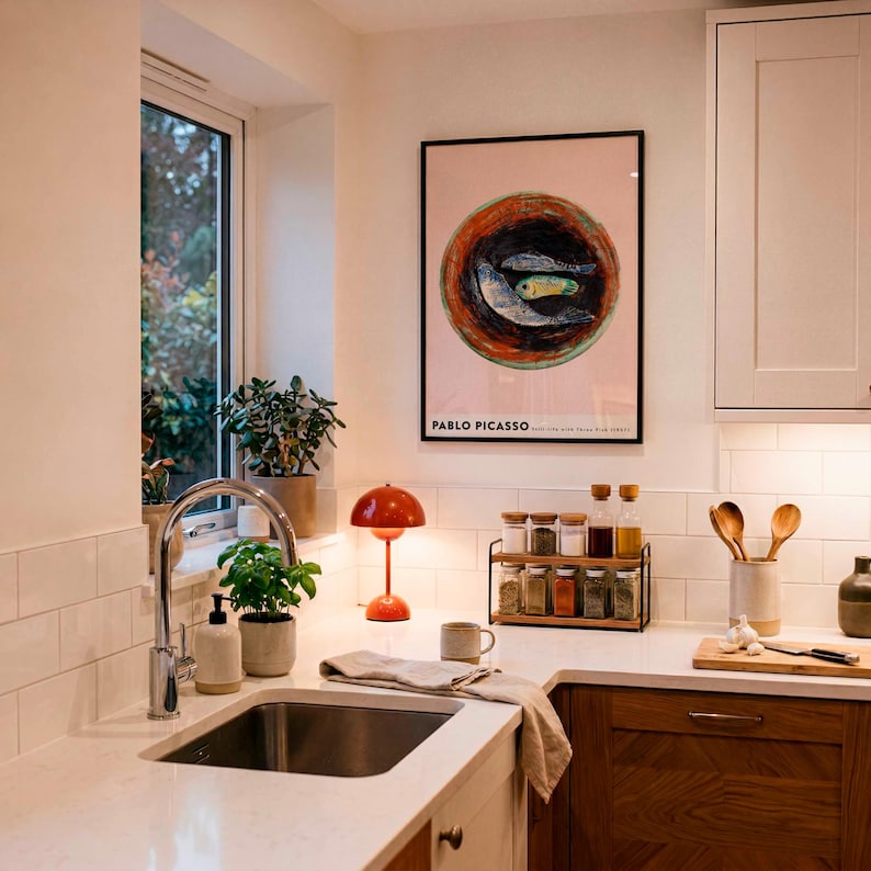 May include: A kitchen scene with a sink, countertop, and cabinets. A framed Pablo Picasso print hangs on the wall. A small orange lamp sits on the counter, along with spice jars, wooden spoons, and a cutting board.