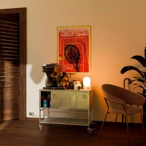 May include: A room scene featuring a coffee station on a rolling cart, a framed tennis poster, and a modern chair. The poster reads "Roland Garros 2002". A small white lamp sits on the cart. The cart has a metal frame and a light green cabinet.