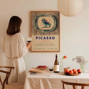 May include: A framed Picasso art print with a blue bird design on a plate, with French text. The print hangs above a table set for a meal, with a bottle of wine, a glass of wine, and a bowl of fruit.