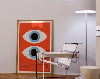 Peut inclure: Une affiche orange Bauhaus avec deux grands yeux bleus, encadrée de bois, est appuyée contre un mur blanc. Une chaise en cuir blanc avec un cadre chromé et un lampadaire gris se trouvent à proximité. Un petit tabouret crème est également présent.