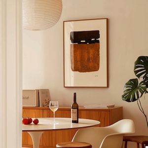 Op de afbeelding: Een abstracte kunstprint in een houten lijst, met een bruin en oranje getextureerd ontwerp. De afbeelding wordt weergegeven in een eetkamer, met een witte tafel, een fles wijn en een glas wijn. Een monstera plant is zichtbaar.