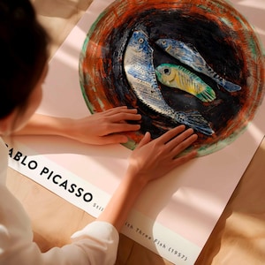 May include: A poster featuring a still life artwork of three fish in a bowl, with the text "Pablo Picasso" and "Still Life with Three Fish (1957)". The fish are blue, yellow, and green, set against a dark background.