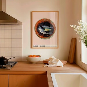 May include: A framed art print of Pablo Picasso's "Still Life with Three Fish (1921)". The artwork features three fish in a bowl with orange and black tones, set against a pink background. The print is in a wooden frame.