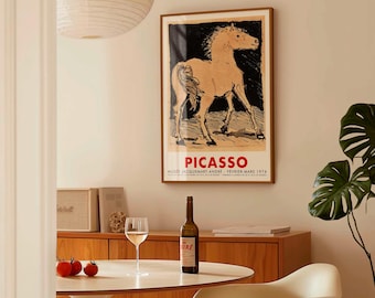 May include: A framed Picasso art print featuring a horse illustration in beige and black tones. The print is displayed above a wooden cabinet. A bottle of wine, a glass of wine, and tomatoes are on a white table.