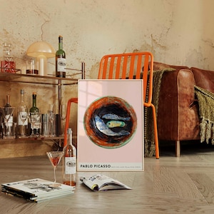 May include: A framed art print featuring a Picasso painting of three fish in a bowl, set against a pink background. The print is displayed on a wooden floor, near a bar cart with bottles and glasses, a martini glass, and a magazine.