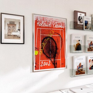 May include: Framed artwork featuring a tennis court design with a red background and the text "Roland Garros 2002". The artwork includes a tennis racket and ball, with a vintage aesthetic. Other framed art and magazines are also visible.