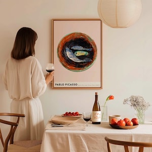 May include: A framed Pablo Picasso print featuring a still life with three fish. The artwork is displayed above a table set for a meal, with a bottle of wine, a glass of red wine, and a plate of tomatoes. A person is holding a glass of red wine.