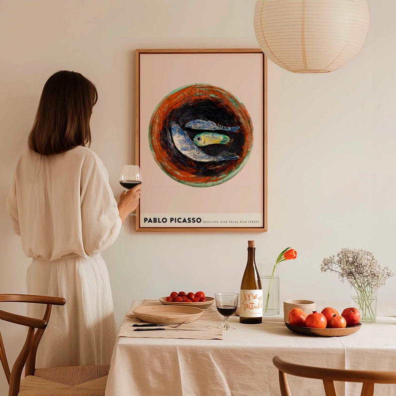 May include: A framed Pablo Picasso print featuring a still life with three fish. The artwork is displayed above a table set for a meal, with a bottle of wine, a glass of red wine, and a plate of tomatoes. A person is holding a glass of red wine.