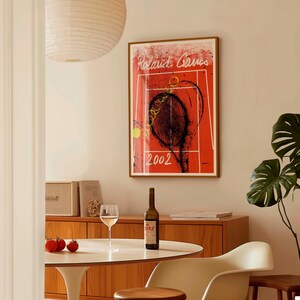 May include: A framed art print featuring a tennis court design with the text "Roland Garros 2002" in a living room setting. A bottle of wine and a glass of wine are on a white table. A wooden cabinet and a plant are also visible.
