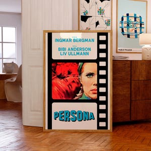 以下が含まれることがあります： イングリッド・バーグマンの映画「ペルソナ」のフレーム入り映画ポスター。ビビ・アンデションとリヴ・ウルマンが出演。ポスターは、映画のタイトルが太字の青い文字で書かれた白黒のフィルムストリップのデザインです。画像には女性の顔と赤い髪が含まれています。