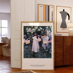 May include: A framed art print of John Singer Sargent's Carnation, Lily, Lily, Rose painting, featuring two children in white dresses holding lanterns in a garden. Above it, a vertical striped poster and a Bialetti coffee maker print are displayed.