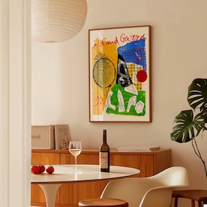 May include: A framed art print with the words "Roland Garros" in red, featuring a tennis racket and abstract shapes in yellow, blue, and green. A bottle of wine and a glass are on a white table. The room has a mid-century modern style.