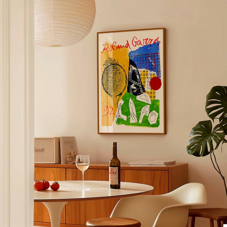 May include: A framed art print with the words "Roland Garros" in red, featuring a tennis racket and abstract shapes in yellow, blue, and green. A bottle of wine and a glass are on a white table. The room has a mid-century modern style.