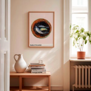 May include: A framed art print featuring a Picasso painting of three fish in a bowl. The artwork has a pink background with a brown and green bowl. The print is in a light brown wooden frame. The text "PABLO PICASSO" is at the bottom.