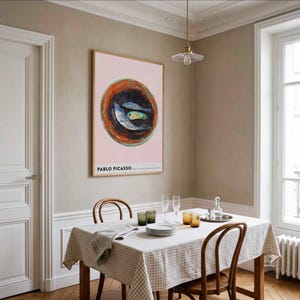May include: A dining room scene featuring a framed Pablo Picasso artwork with a pink background and a fish illustration. The table is set with plates, glasses, and a checkered tablecloth. Wooden chairs and a hanging light fixture complete the decor.