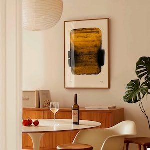 May include: A framed abstract art print with a yellow and black rectangular design hangs on a beige wall. A bottle of wine, a wine glass, and tomatoes sit on a white table. A wooden cabinet and a plant are also visible.