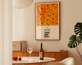 Può includere: Un poster incorniciato con un design astratto arancione e nero è appeso a una parete. Una bottiglia di vino, un bicchiere da vino e tre pomodori sono su un tavolo bianco. Sono visibili anche un mobile in legno e una pianta di monstera.