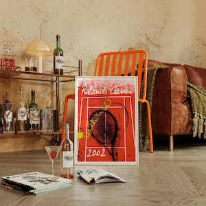 May include: A framed red tennis art print with the words "Roland Garros" and "2002". The image also shows a bar cart with bottles, glasses, and a lamp, an orange chair, and a brown leather sofa.
