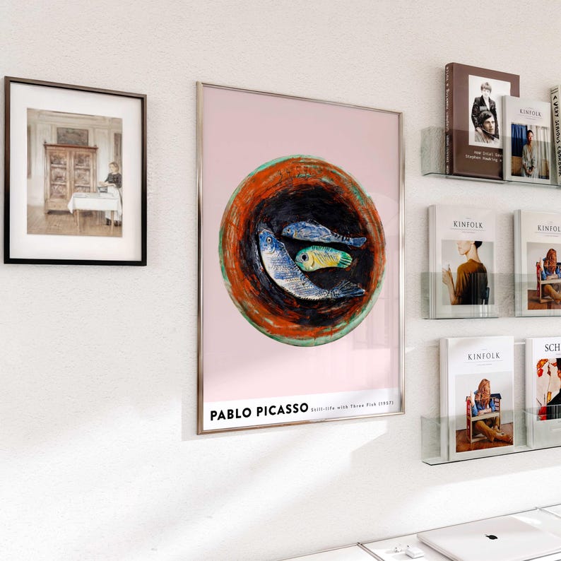 May include: Framed art print of a still life with three fish by Pablo Picasso. The artwork features three fish in a bowl with a pink background. A framed black and white photograph hangs to the left. Magazines and books are displayed on the right.