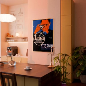 May include: A framed vintage Leica camera advertisement featuring a man holding a camera, with the Leica logo and text in German. The poster has a blue background with orange and black accents, displayed in a cafe setting.