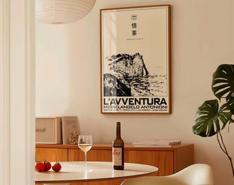 May include: A framed movie poster for "L'Avventura" with Japanese text and a black and white image of a rocky coastline. A bottle of wine and a glass of wine sit on a white table. Three red tomatoes are also on the table.