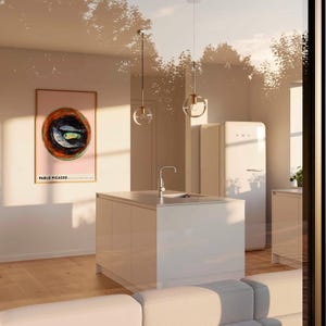 May include: A modern kitchen with a white island, a white refrigerator, and two pendant lights with glass globes. A framed Pablo Picasso artwork hangs on the wall. The room has hardwood floors and is bathed in sunlight.