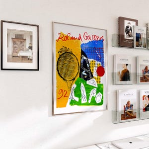 May include: A framed art print with a tennis racket design, the text "Roland Garros", and the number "92". The artwork uses yellow, blue, and green. A framed black and white photograph is to the left. Magazines are on the right.