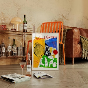 May include: A framed art print with the words "Roland Garros" in red, featuring a tennis racket and court design in yellow, blue, and green. The image is propped up against an orange chair, with a bar cart and bottles in the background.