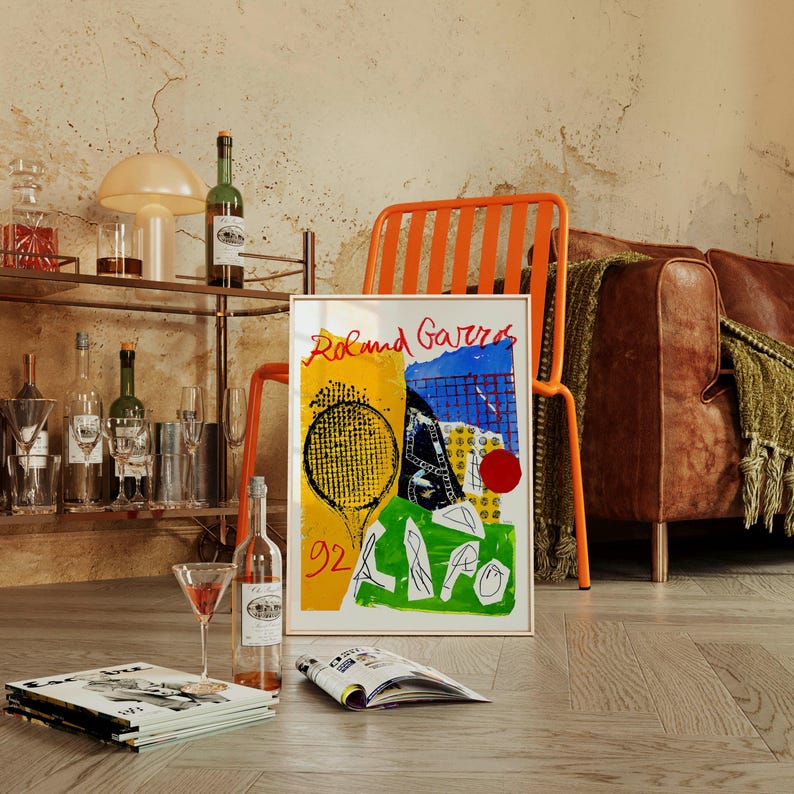 May include: A framed art print with the words "Roland Garros" in red, featuring a tennis racket and court design in yellow, blue, and green. The image is propped up against an orange chair, with a bar cart and bottles in the background.