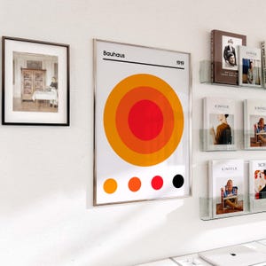May include: A framed Bauhaus art print featuring concentric orange and red circles, with the word "Bauhaus" and the year "1919" printed at the top. A framed black and white photograph hangs to the left. Magazines and books are displayed on the right.