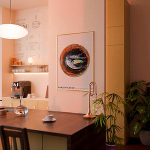 May include: A framed artwork featuring a circular abstract design with a fish in the center, attributed to Pablo Picasso. The artwork is displayed in a cafe setting, alongside a trumpet and a plant.