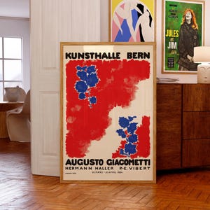 May include: A framed poster with the text "KUNSTHALLE BERN" in black letters. The poster features a red and white background with blue floral designs. Additional posters are visible in the background, including one with the text "JULES et JIM".