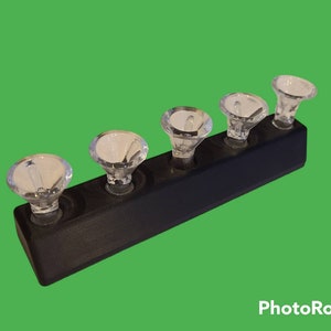 Puede incluir: Un expositor de madera negra con cinco pomos de cristal transparente facetado. Los pomos están espaciados uniformemente en la parte superior del expositor. El expositor es rectangular con un frente ligeramente inclinado. Aproximadamente 20 cm de largo.