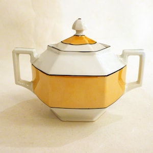 Peut inclure: Un sucrier vintage au design géométrique. Le sucrier à couvercle présente une palette de couleurs blanche et jaune avec des bordures noires. Il possède deux poignées et un petit bouton sur le couvercle. Le sucrier est probablement en céramique.