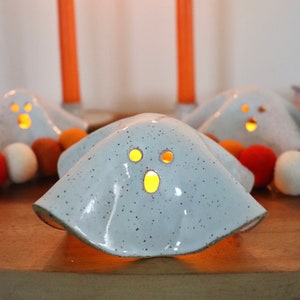 May include: Ceramic ghost-shaped candle holders with speckled gray finish and cut-out eyes and mouth, emitting a warm glow. Decorative Halloween-themed items with orange and white felt ball accents and orange candles in the background.