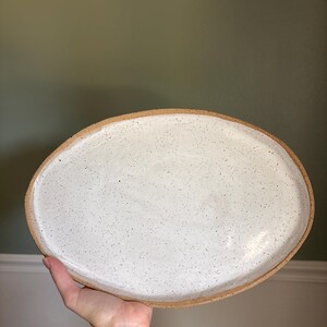 May include: An oval-shaped ceramic platter featuring a speckled white glaze on the interior surface and a raw, unglazed tan clay rim. The plate has a smooth, slightly irregular edge and a flat base, showcasing a natural, earthy aesthetic.