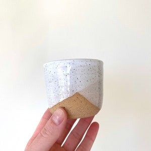 May include: A small, handmade ceramic cup with a speckled white upper half and a tan, triangular-shaped base. The cup is held in a hand against a plain white background. The cup is likely used for drinking.