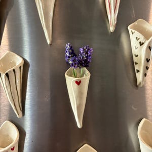 May include: A collection of cone-shaped ceramic wall vases in off-white, each featuring unique hand-painted designs including small red and blue hearts, black stripes, and patterns. One central vase holds two sprigs of purple lavender flowers.